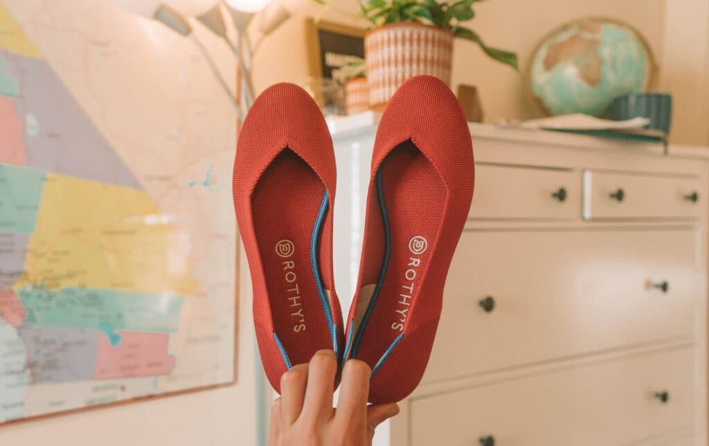 A hand holds a pair of red Rothy's flats up with a soft-focus white dresser and bedroom in the background.