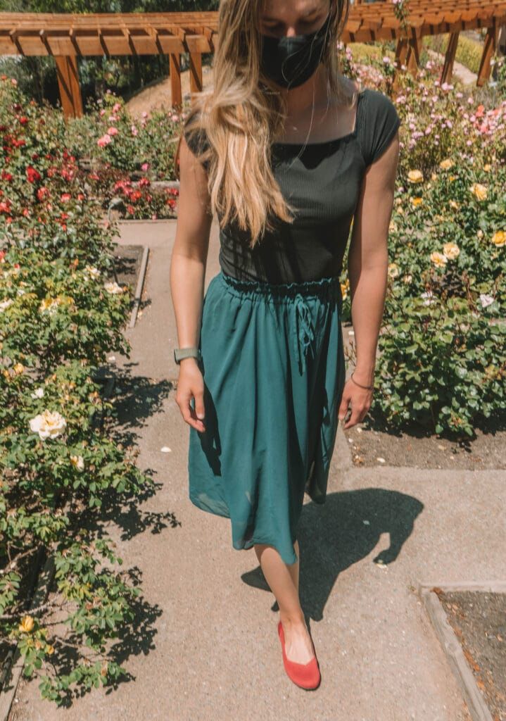 A light-haired woman wearing a turquoise skirt, a black face mask, and a pair of red Rothy's flats walks on a path through a lushly blooming rose garden.