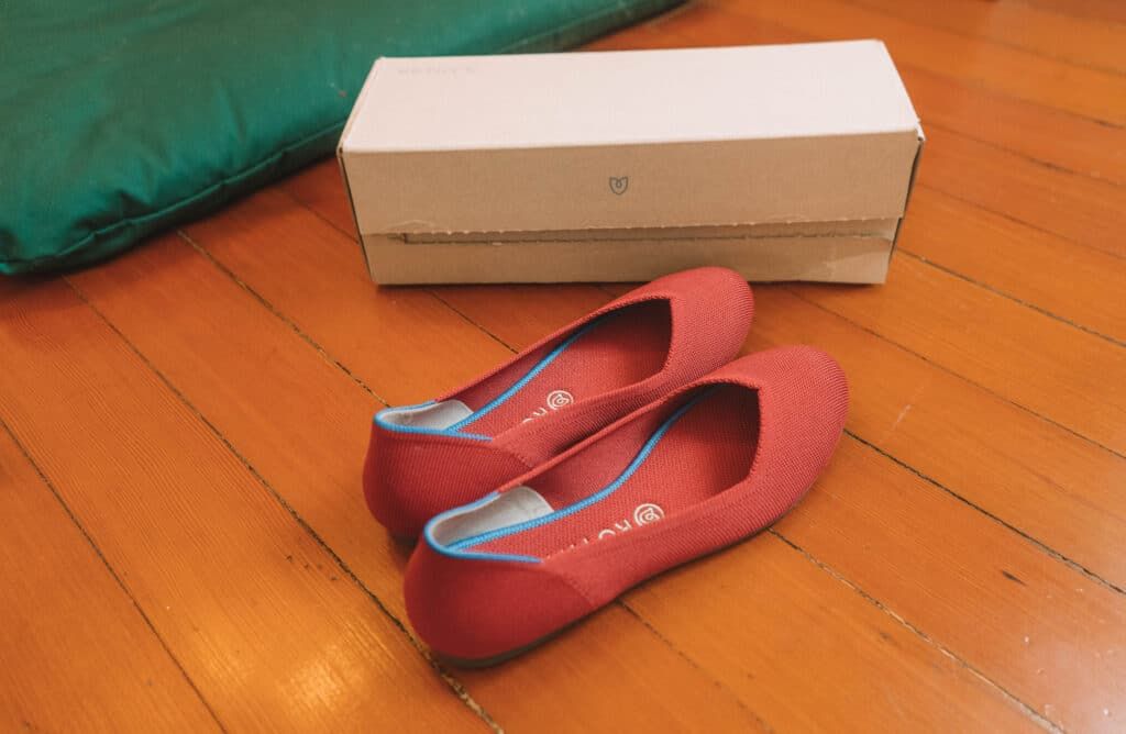 A pair of red Rothy's flats sitting on a hardwood floor with a shoebox behind them.