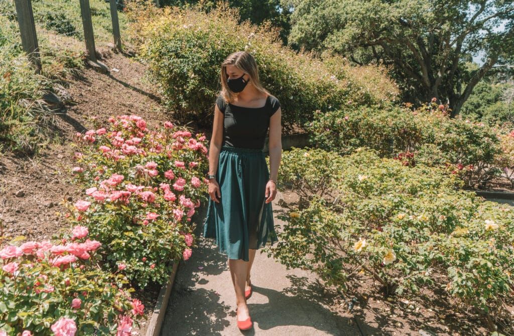 A light-haired woman wearing a turquoise skirt, a black face mask, and a pair of red Rothy's flats walks on a path through a lushly blooming rose garden with trees in the background.