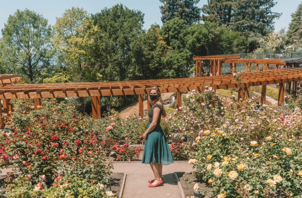 A light-haired woman wearing a turquoise skirt, a black face mask, and a pair of red Rothy's flats stands on a path through a lushly blooming rose garden.