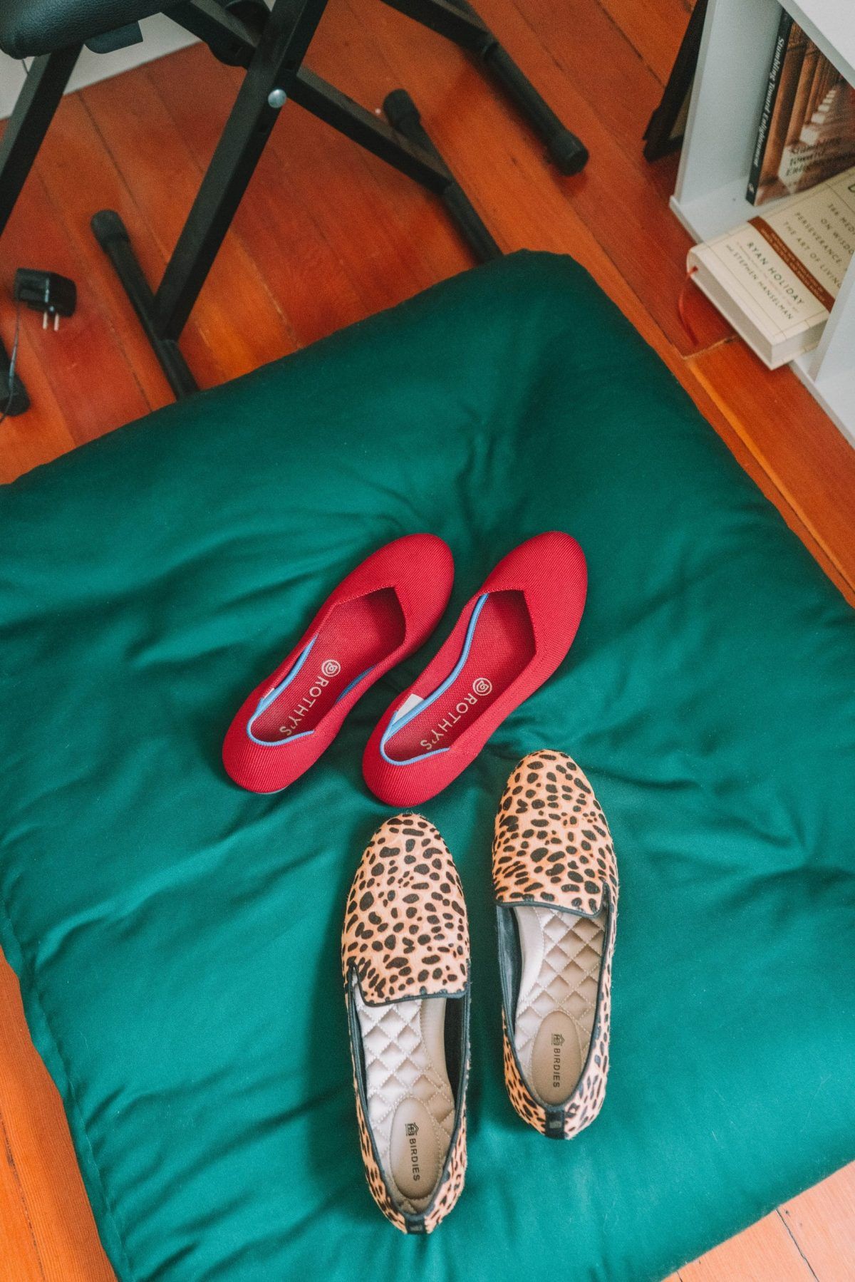A photo comparing Rothys vs Birdies shoes, with a pair of red Rothys flats and a pair of cheetah-print Birdies flats sitting on a dark green pillow.