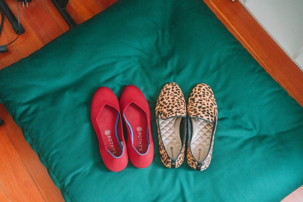 A pair of red Rothys flats and a pair of cheetah-print Birdies flats sitting on a dark green pillow.