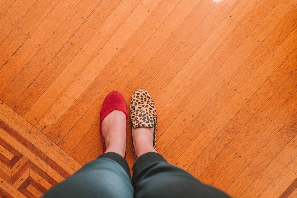 POV-shot of a woman's feet wearing one red Rothys flat and a cheetah-print Birdies flat standing on a hardwood floor with an inlaid wood parquet border.