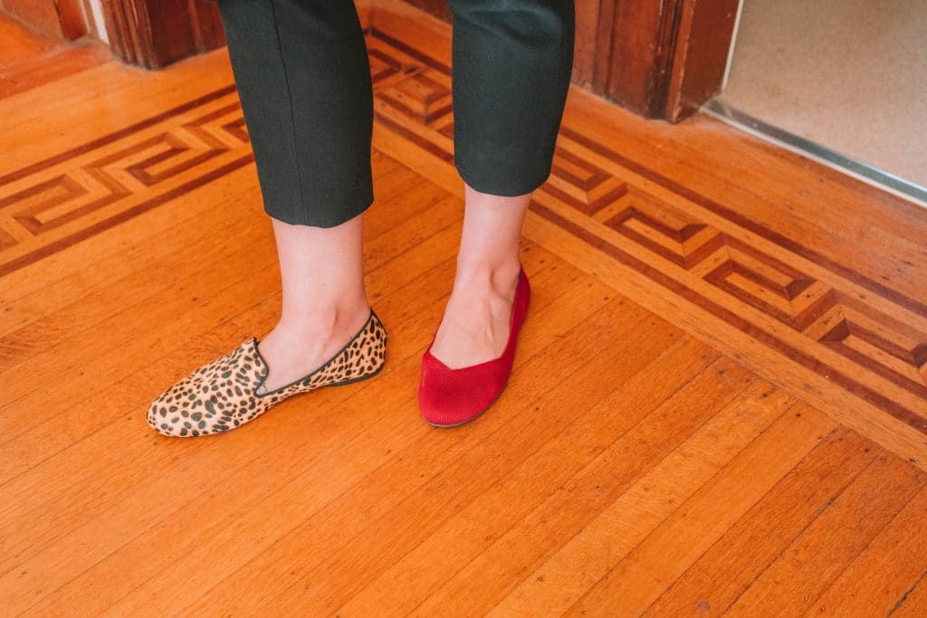 A woman's feet comparing rothys and birdies shoes side by side by wearing on red Rothys flat and a cheetah-print Birdies flat standing on a hardwood floor with an inlaid wood parquet border.