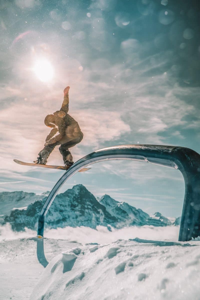 Snowboarder grinding at a snow park with the sun, mountains, and snow behind them.