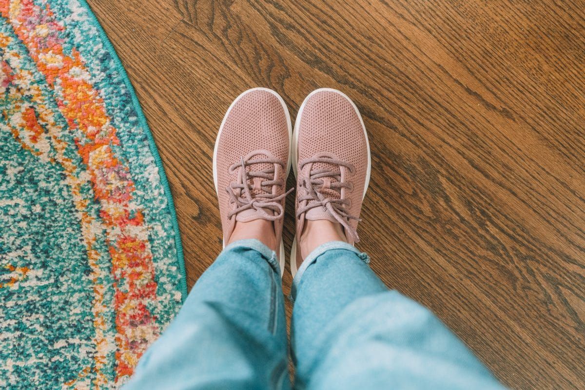 A POV-view looking down at a pair of feet wearing pink Allbirds standing on a hardwood floor next to a colorful rug.