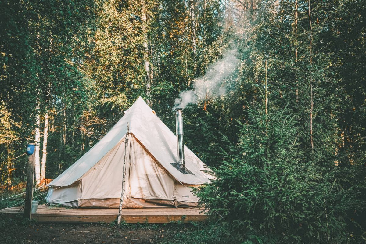 A wide shot of a canvas bell tent on a wooden platform with a chimney from a tent stove emitting smoke from the roof and thick evergreen trees in the background. 