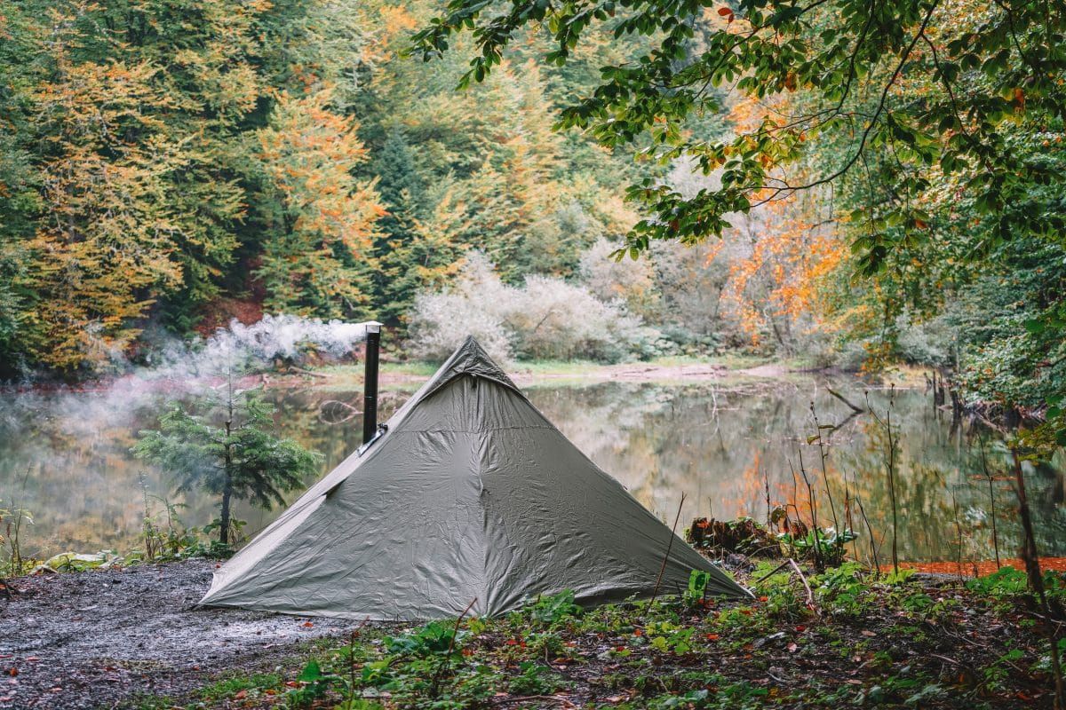 A small, dark green, teepee-style hot tent with smoke coming from its chimney, and a lake and autumn leaves in the background.