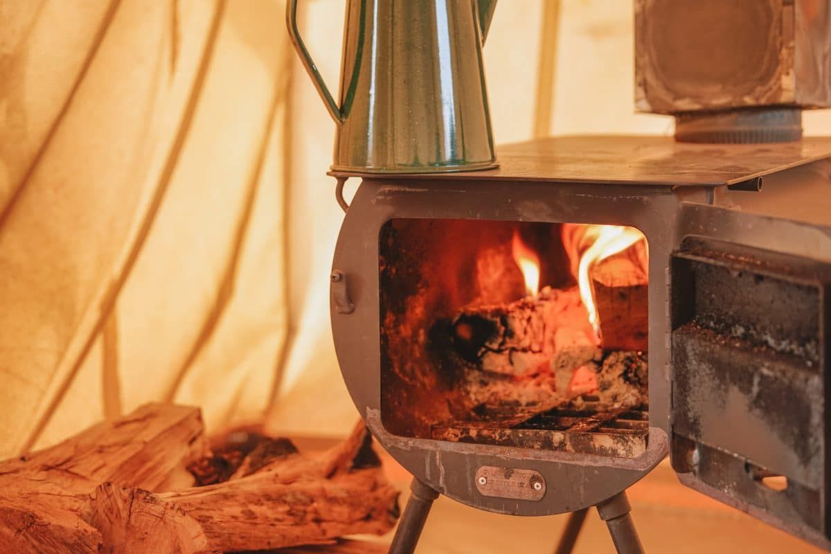 A close-up image of a wood fire inside a cast iron tent stove in a canvas tent, with a metal kettle sitting on top of it.
