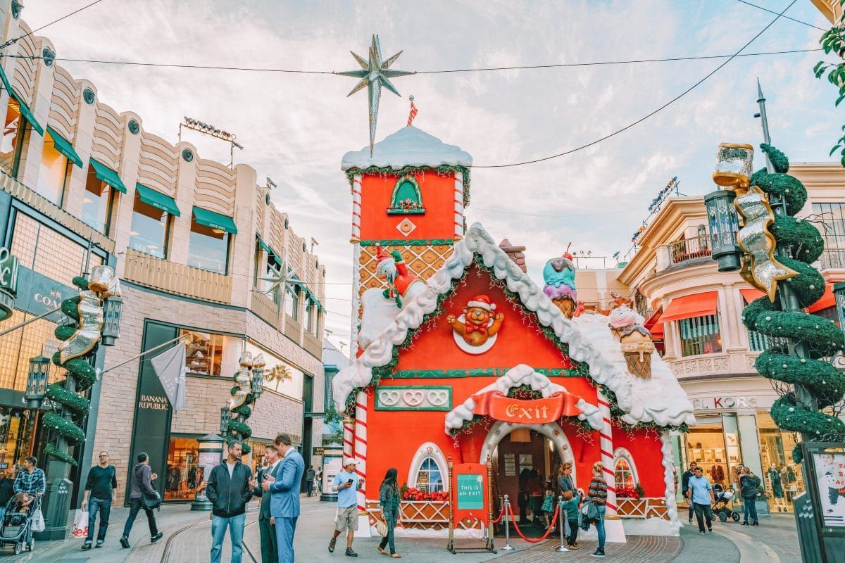 Sparse crowds of people stroll past a red, amusement park-style, life-sized gingerbread house in an outdoor shopping center.