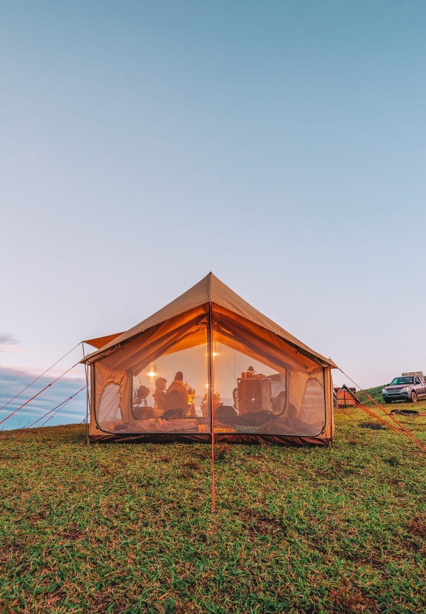 A example of one of the best screen houses for camping: a large, a-frame canvas screen house illuminated at dusk on a grassy campsite overlooking the sea.