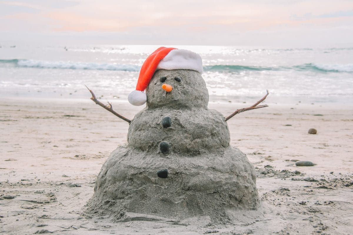 A 'snowman' made from sand on the beach, complete with a carrot nose and a Santa hat.