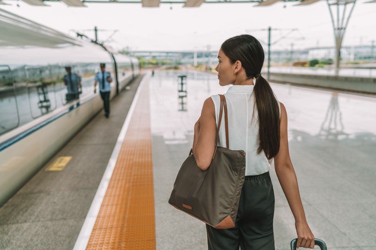 A woman with a long dark ponytail seen from behind as she walks down a train platform, pulling the handle of rolling luggage, with a large purse on  her shoulder.