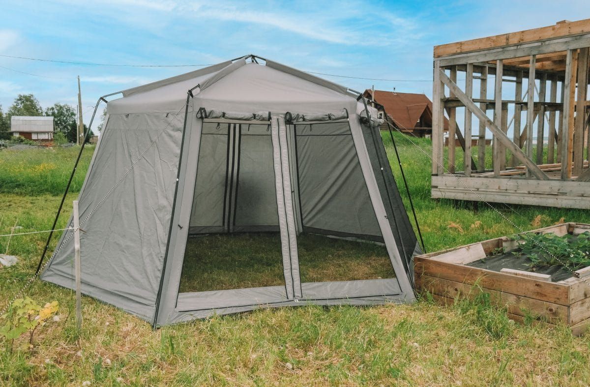 A grey screen house tent pitched in a garden next to a wooden deck and flower bed, with a blue sky in the background.