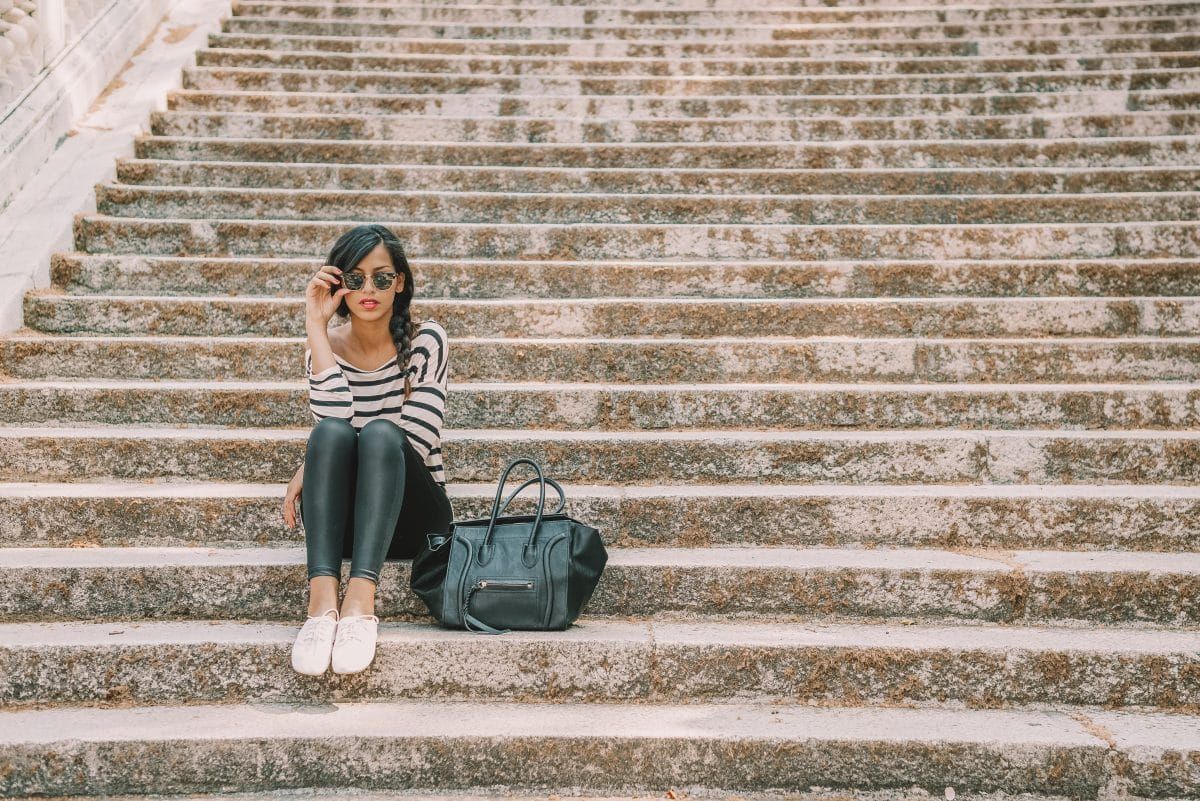 A stylish woman wearing sunglasses, black leggings and a striped shirt sits on stone steps with a black leather purse beside her.