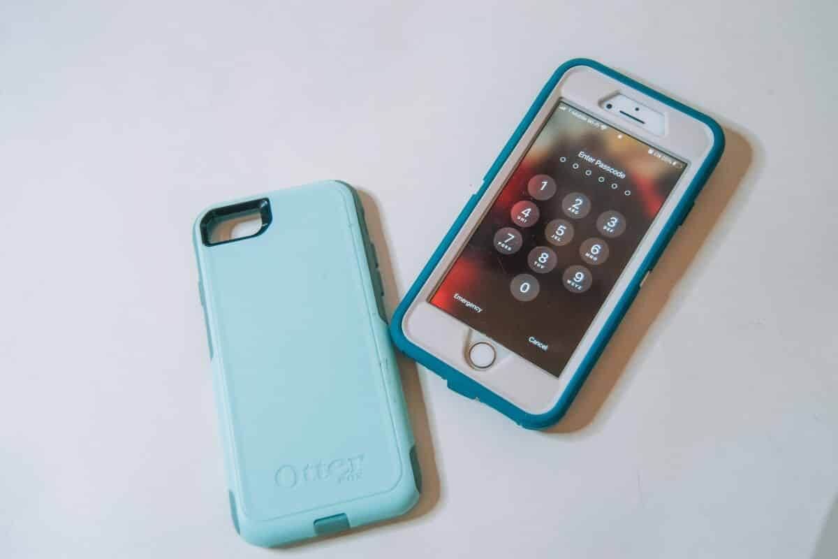 An iPhone in a dark teal Otterbox Defender phone case, sitting on a white background with the home screen facing up, beside a light teal commuter case.