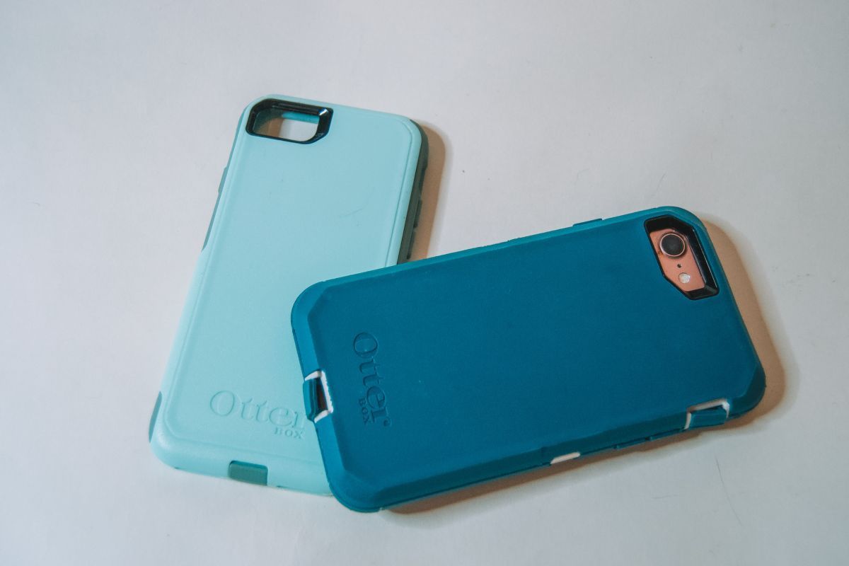 An iPhone in a dark teal Otterbox Defender phone case, sitting on a white background with the home screen facing down, beside a light teal commuter case.