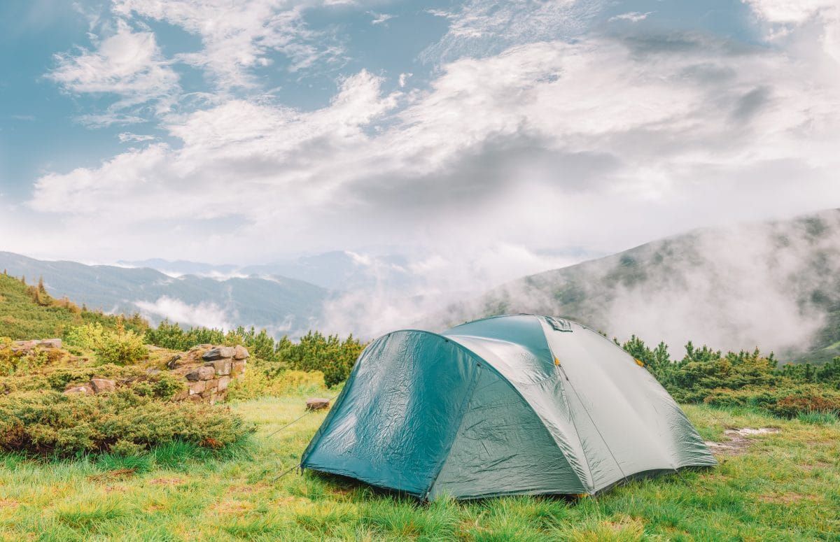 A wet-looking, dark green-blue tent pitched in a grassy campsite, with mist rising from the valley behind it, and sun coming out from behind the clouds.