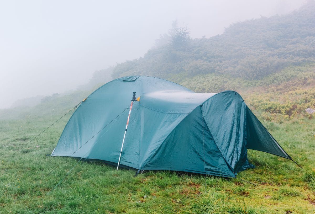 A wet-looking, dark green-blue tent pitched in a grassy, misty campsite.