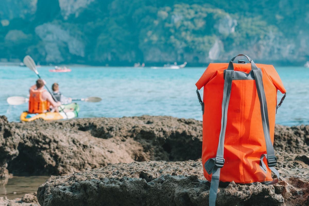 An orange dry bag sitting on volcanic rocks, with a couple in a tandem kayak paddling on the sea in the background.