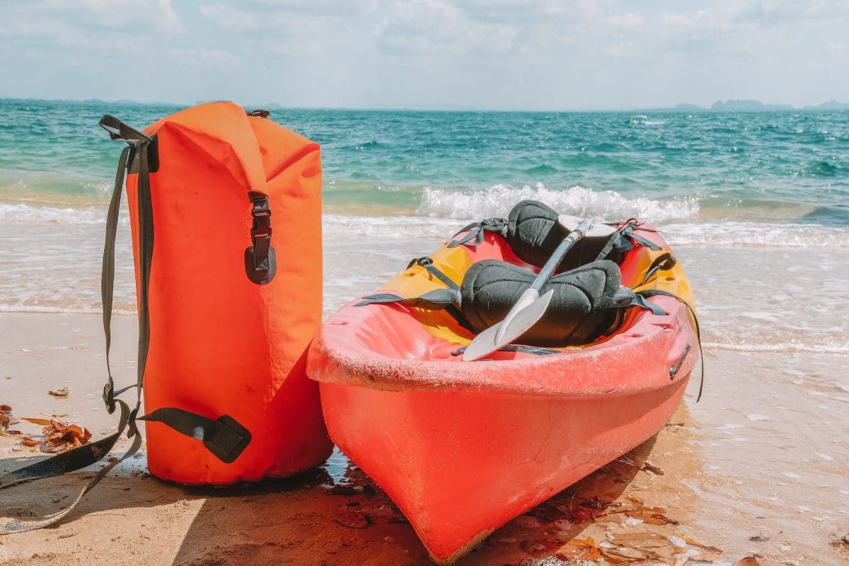 An orange kayak sitting next to an orange dry bag on a sandy beach, with turquoise ocean water in the background.