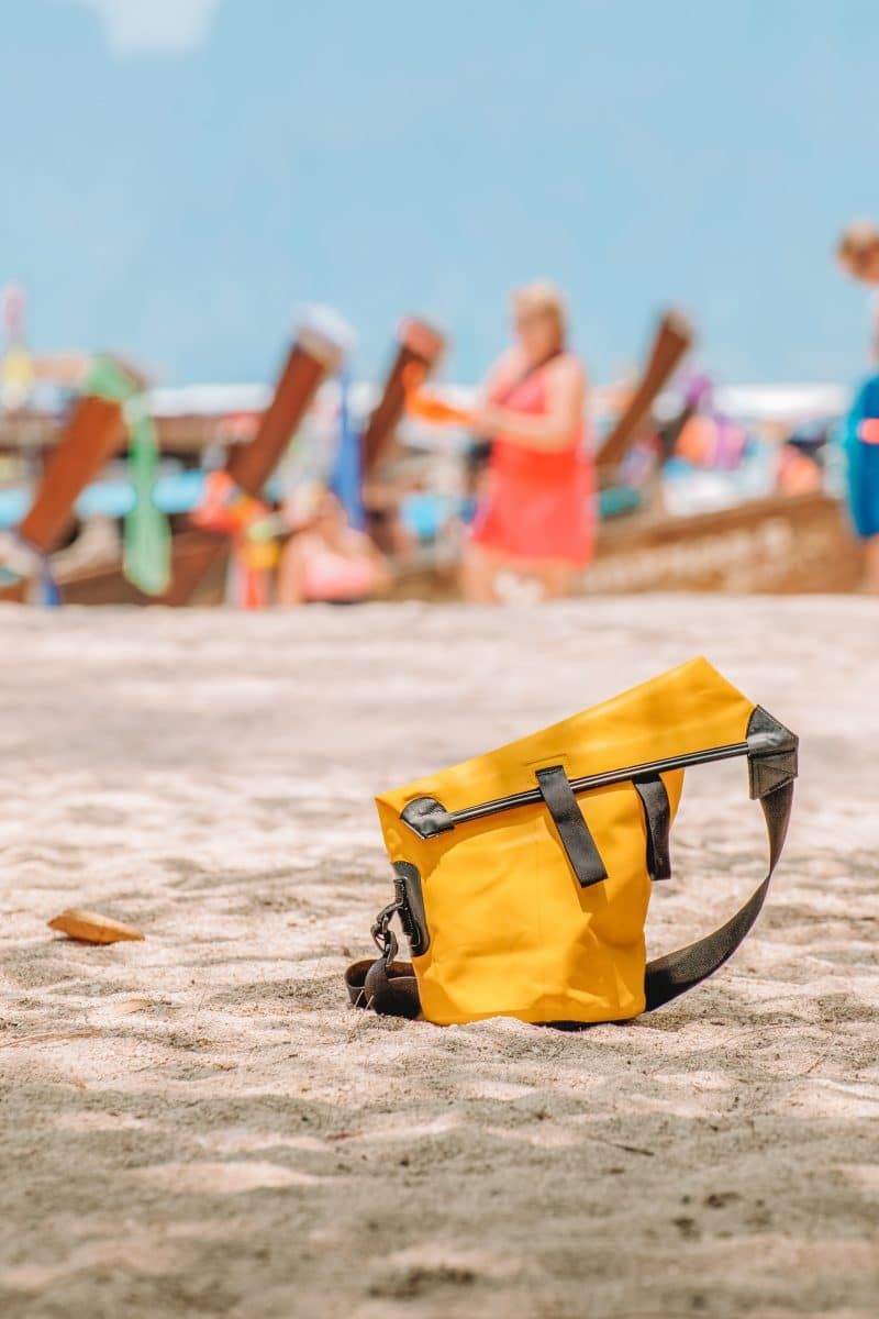 A yellow kayaking dry bag sitting on a sandy beach with colorful boats in soft focus in the background.