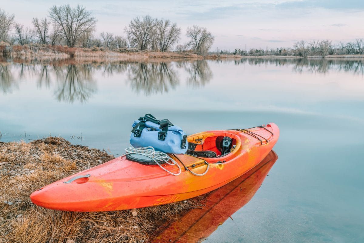 An orange kayak banked on the side of a lake with a blue dry bag balanced on top of it, an overcast sky and wintry trees in the background.