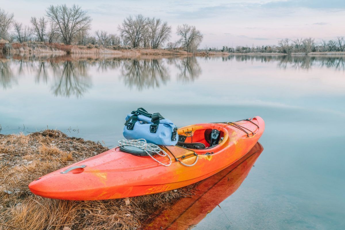 An orange kayak sits at the edge of the water, with the stern grounded on land. A blue waterproof duffle bag and rope sit on top.