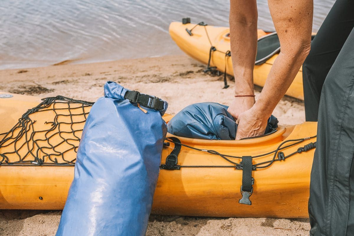 A blue dry bag leans against a yellow kayak on a sandy shore, with a pair of arms visible behind, packing a second dry bag into the vessel.
