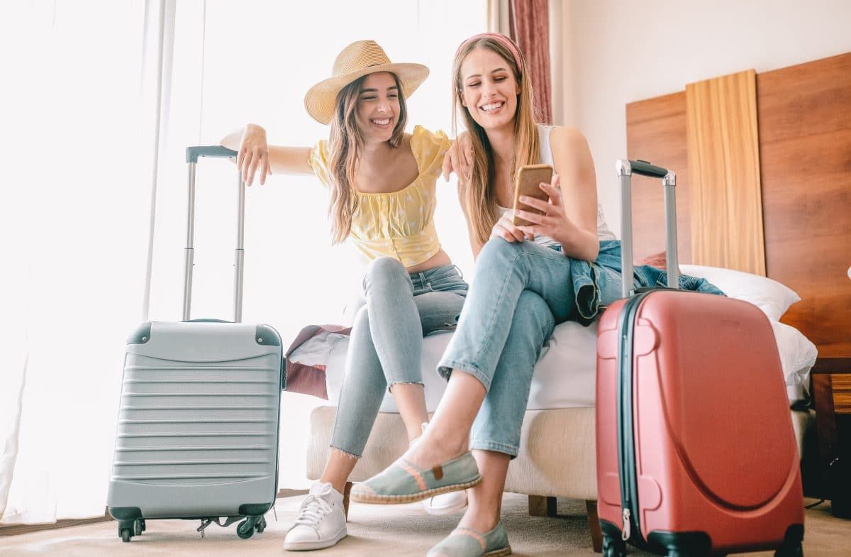 Two young women wearing jeans smile as one shows the other something on her phone as they sit side-by-side on a hotel bed, each with a hardside suitcase beside them.