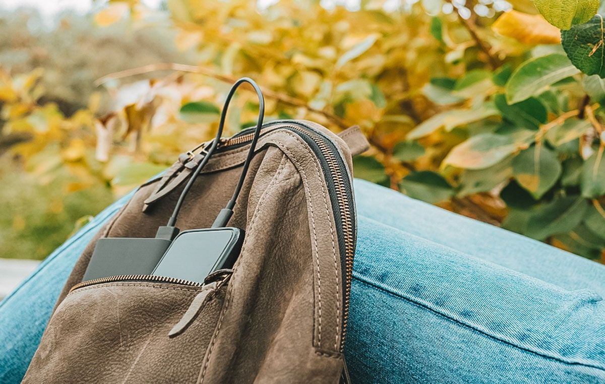 A close-up of a brown leather backpack with a phone and portable charger visible in the front pocket leans against a blue jeans-clad leg with green foliage in the background.