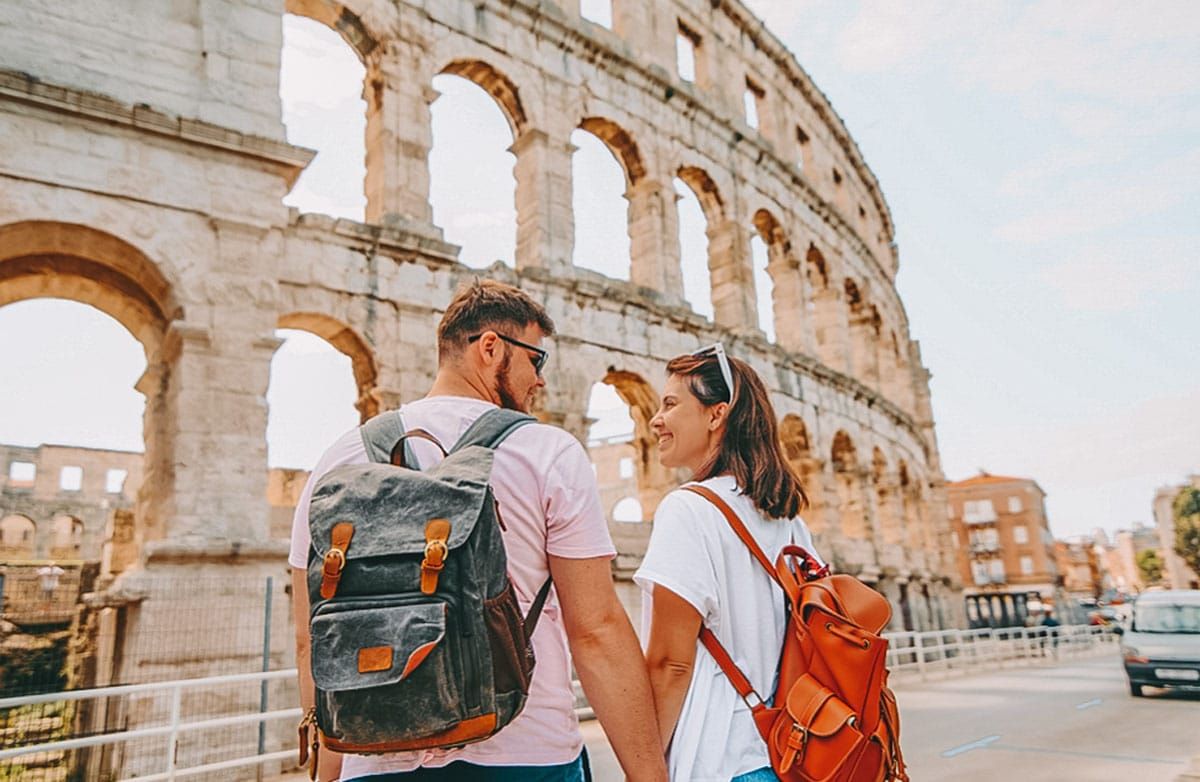 A man wearing a pink t-shirt and a grey backpack and woman wearing a white t-shirt and a brown leather backpack hold hands, while looking at each other and smiling with The Coliseum in the background.