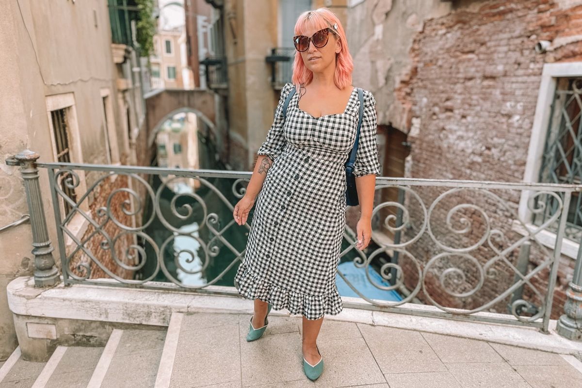 A woman with pink hair, sunglasses, and a black-and-white checkered dress wears a pair of teal Vivaia flats on a bridge over a Venetian canal.