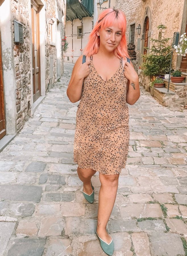 A woman with pink hair and a cheetah-print dress wearing a pair of teal Vivaia flats and walking down a cobblestone street.