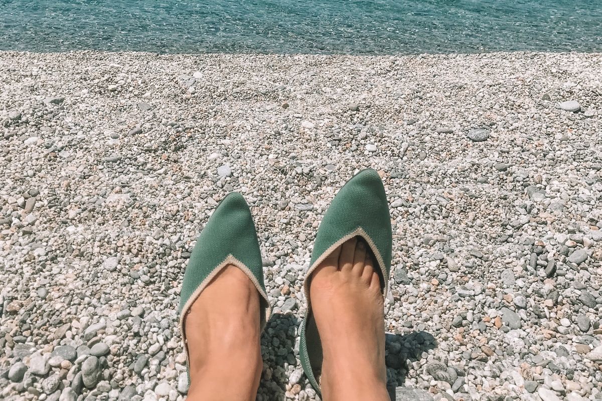 POV shot of a pair of feet wearing a pair of teal Vivaia flats on a pebbled beach with aqua water in the background.