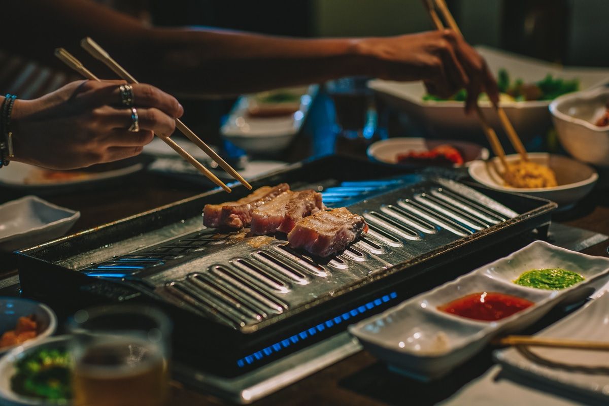 A hand holding chopsticks, poised to pluck a piece of meat from a hibachi grill at a Korean BBQ restaurant.
