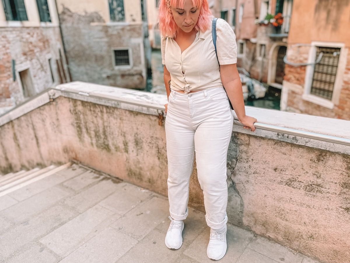 A woman with pink hair wearing a white blouse, white jeans, and white Vessi shoes stands on a bridge in Venice looking down at her feet.