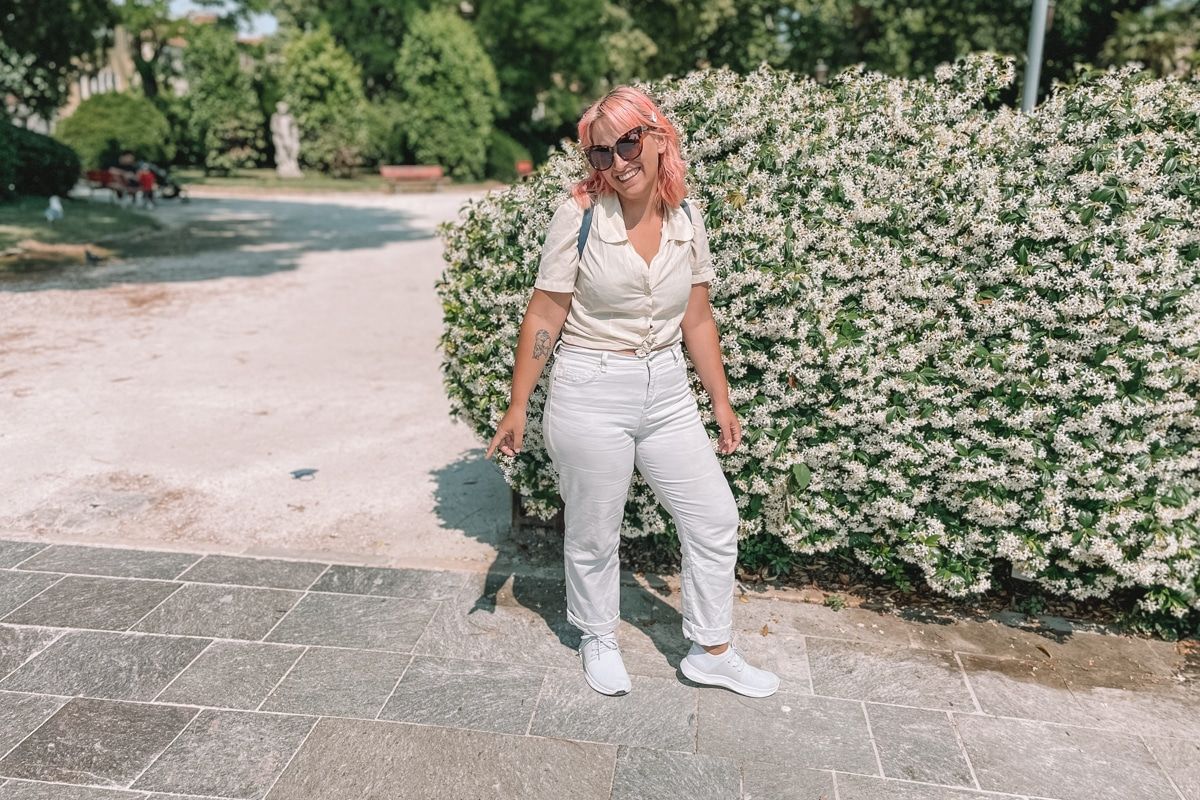 A woman with pink hair wearing sunglasses, a white blouse, white jeans, and white Vessi shoes in front of a bush covering in white flowers, laughing a the camera.
