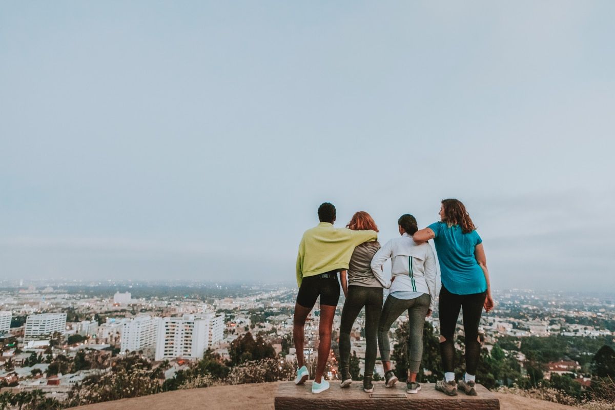 A group of four women in exercise clothes stand on the top of a hill looking out over the city.