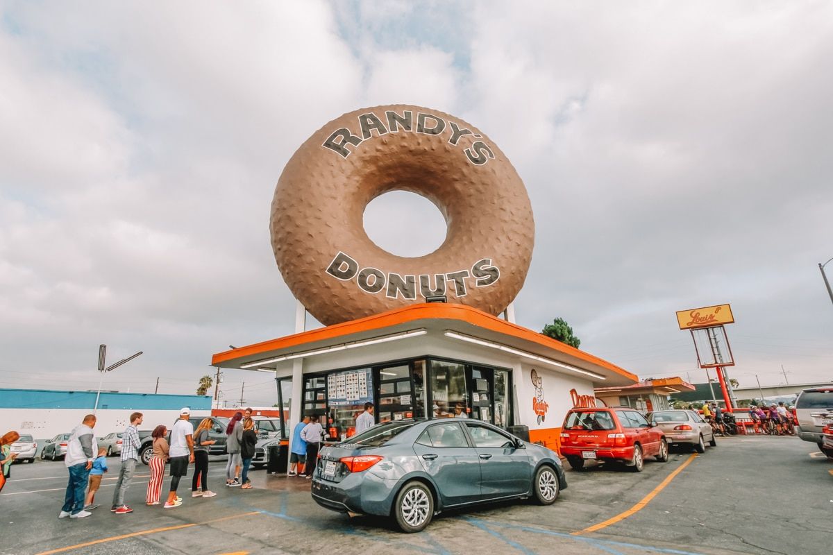 Cars and people waiting outside Randy's Donuts, a small stand with a giant model donut on the roof.