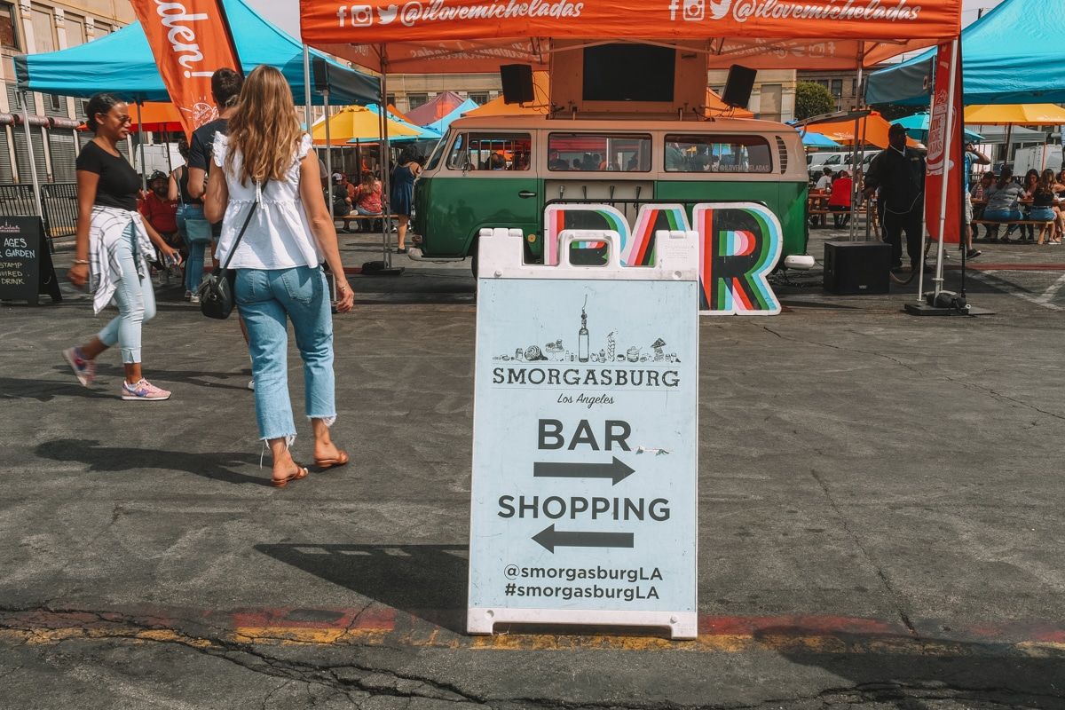 People milling around a sandwich board sign advertising Smorgasburg LA, with tented stands in the background.