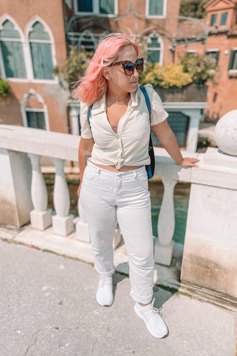 An honest review of Vessi shoes: a woman with pink hair wearing sunglasses, a white blouse, white jeans, and white Vessi shoes stands on a bridge in Venice looking over her shoulder.