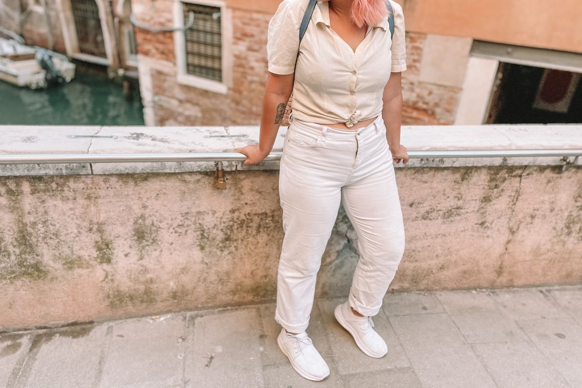 A woman with pink hair wearing a white blouse, white jeans, and white Vessi shoes seen from the neck down, standing on a bridge in Venice.