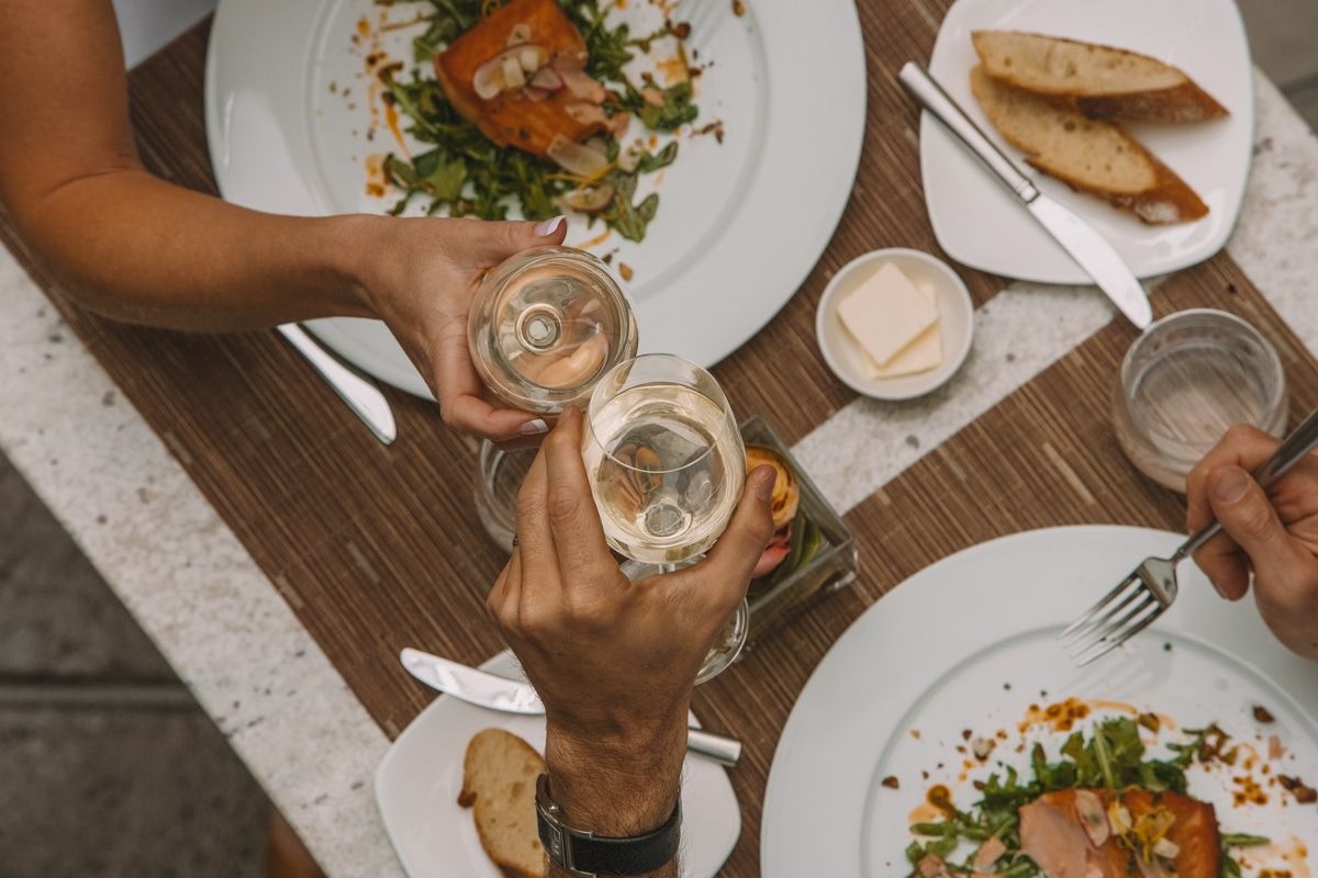 Bird's eye view of a restaurant table with plates of food and two hands toasting with glasses of white wine.