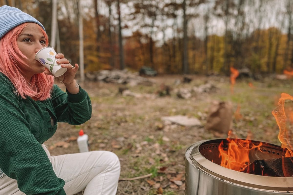 Woman with pink hair wearing a green shirt, sipping a dixie cup while sitting next to roaring fire in a Solo Stove.
