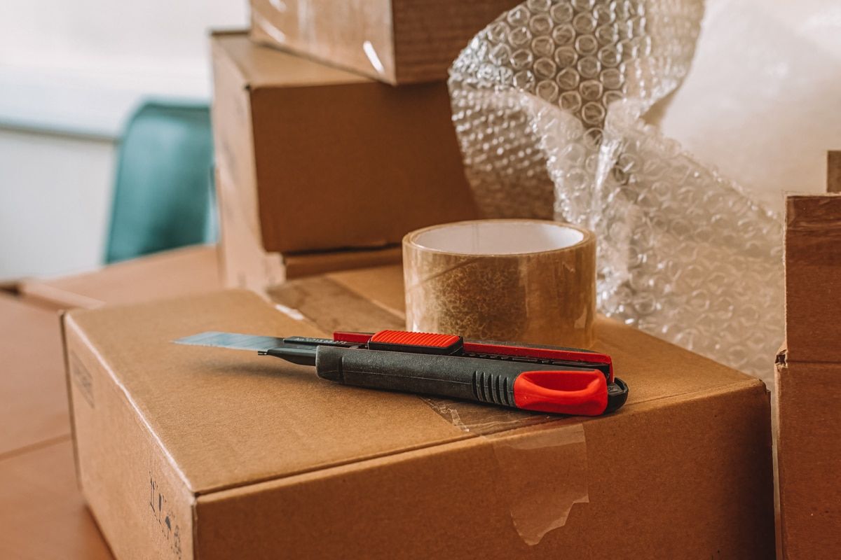 Packaging materials such as bubble wrap, tape, and box-cutter sitting on top of cardboard box.