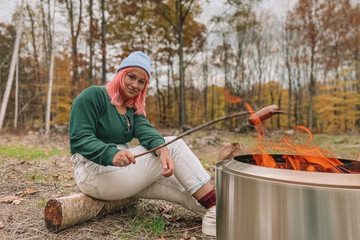 Woman with pink hair in a green shirt roasting a sausage over a Solo Stove bonfire with a forest and fall leaves in the background.