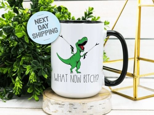Coffee mug printed with a green t-rex and the phrase, "What now, Bitch?