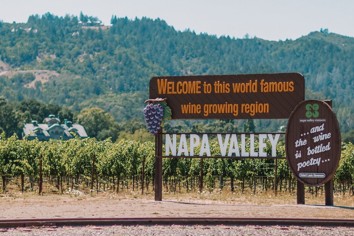 A sign reading "Welcome to this World Famous Wine Growing Region, Napa Valley," with vineyards in the background.
